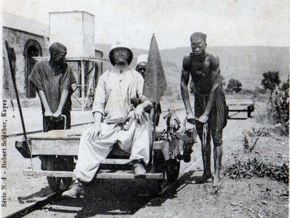 Svart-hvitt foto fra 1904 av franskmann i tropehjelm sittende på en jernbanetralle, dyttet av to afrikanske arbeidere. 